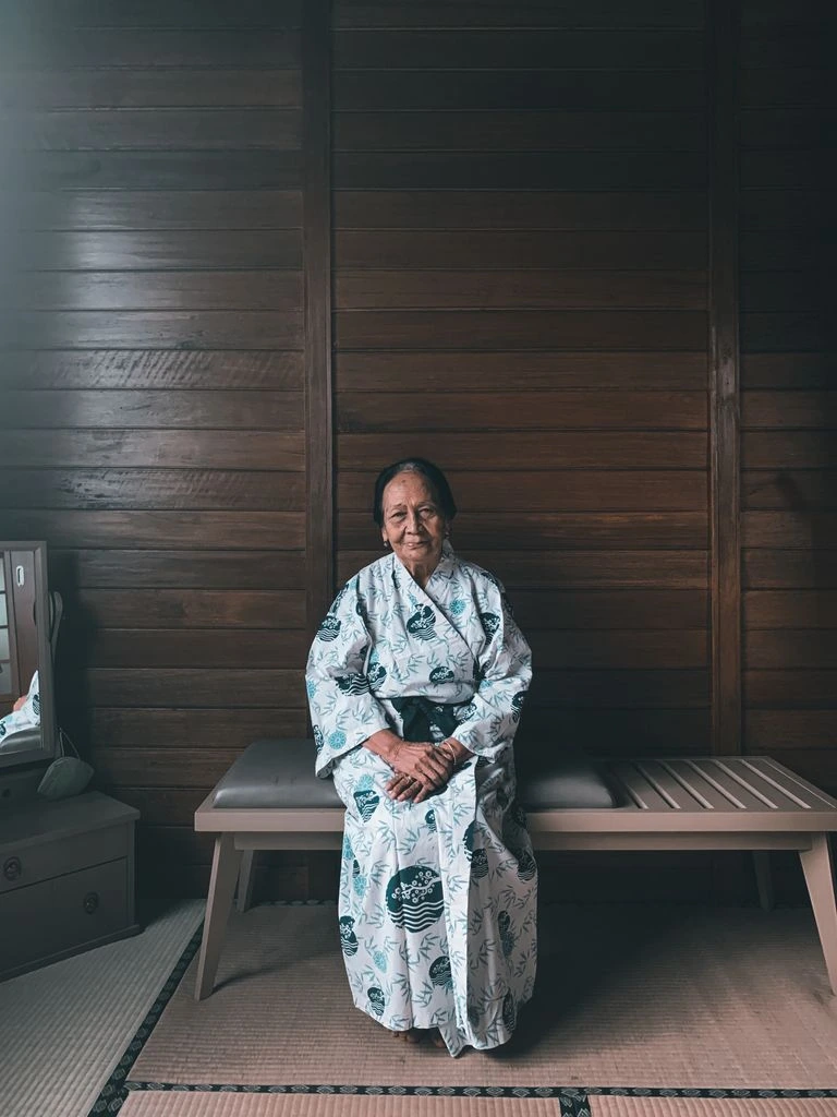 Restored still of an elderly Japanese woman seated in a softly lit kitchen.